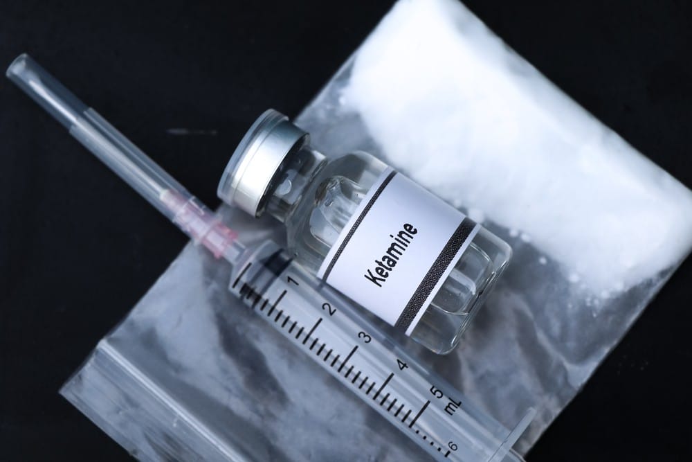 A vial of Ketamine and a syringe next to it with a small bag containing white powder.